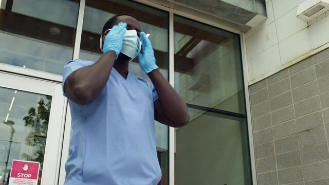 African American Black Male Nurse Doctor Wearing PPE Adjusting Medical Mask And Gloves Walks Out Of Frame, Low Angle Medium Frame Exterior Day. 4K.