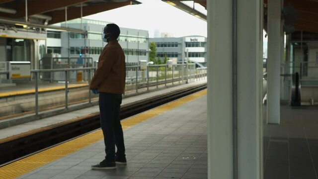 African American black man waiting for train on exterior platform wearing causal clothes and PPE mask and gloves, wide frame camera tracks right to left from behind post. 4K.