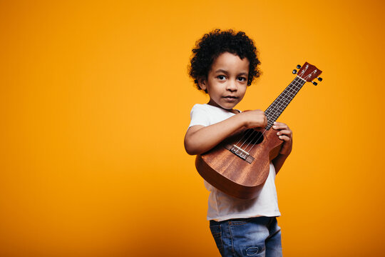 Black-skinned curly boy in a white T-shirt plays ukulele guitar and looks at the camera on an orange background in the studio