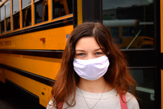 Child Wearing Face Mask By School Bus. A Girl With A Facemask Stands By A School Bus. Education, Medical, Health, Safety, Back To School Concepts.