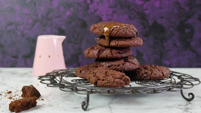Pouring extra chocolate sauce over stack of chocolate chip homemade cookies, in creative vintage setting against a purple and white marble background. Video footage,