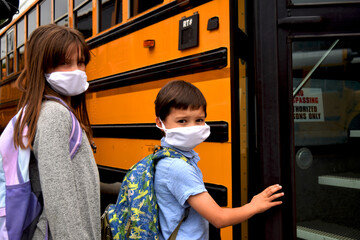 Children wearing face masks by school bus. A boy and girl wear facemasks while boarding a school bus. Education, back to school, school, medical, health, and safety concepts.