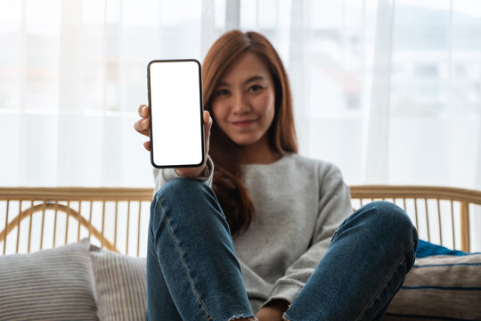 Mockup image of a beautiful asian woman holding and showing a mobile phone with blank white screen while sitting on sofa at home