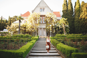 Young beautiful woman, wearing skirt, top and hat, walking in a blooming park among the greenery and the old palace with wisteria.