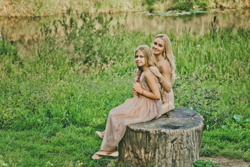 Two adorable blonde girls sitting near the lake on summer sunny day. Outdoor photo of happy sisters posing on nature background. 