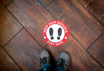 Floor sign on wood floor at business stating Social Distancing Stay Safe with shoes as graphic . A...