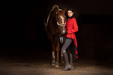 Smiling jockey woman standing with brown horse in stable, black background