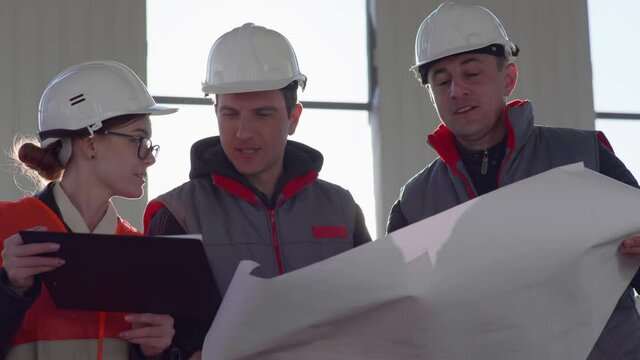 working team of male and female builders in protective helmets with drawings in their hands discuss construction of a covered building, background of a window and sunlight