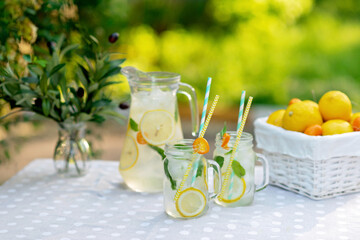 Lemonade refreshing drink in a jug and jars with lemons, fresh mint and ice with a basket with lemons and kumquat on a garden table. Summer outdoor picnic. Soft selective focus.