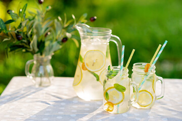 Fototapeta premium Lemonade refreshing drink in a jug and jars with lemons, fresh mint and ice on a garden table. Summer picnic outdoors. Copy space. Soft selective focus.