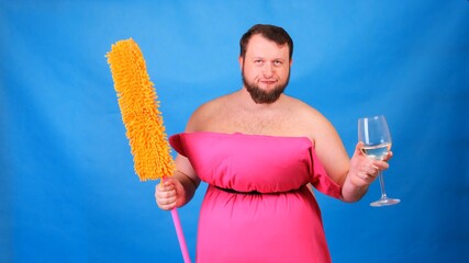 Funny bearded guy in a pink pillow dress with a mop and a glass of wine on a blue background. Crazy quarantine. Funny house cleaning. Fashion 2020. Put on a pillow. Challenge 2020 due to house