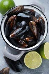 Close-up of raw iced mussels in a grey pan with limes, studio shot on a light-blue stone surface, elevated view