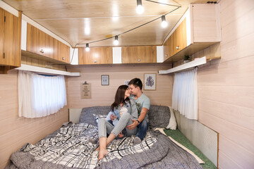 Obraz premium Happy newlyweds hug and kiss inside the trailer track on the couch. A honeymoon trip across the country. Budget vacation for a young family. Beautiful couple relaxing in the camping. Concept Caravan