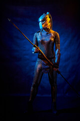 Valkyrie girl looking like an alien in shiny military armor, a strange helmet and a spear in a dark room with plants and vines. Model during a photo shoot, the actress during the shooting of the film.