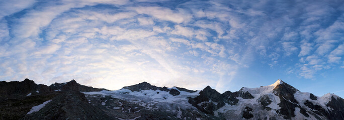 Fototapeta na wymiar Panoramic snapshot of a ridge of rocky mountains in Switzerland in the morning, blue sky with wavy clouds is over snowy peaks, rising sun shining at Ober Gabelhorn