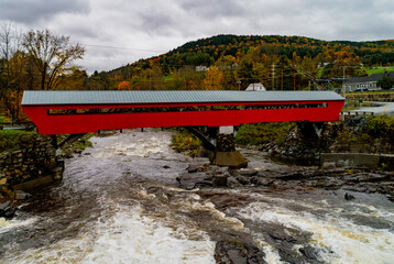 A red covered bridge first built in 1883 spans a rapidly flowing river in Vermont