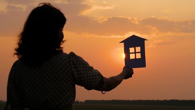 The girl keeps a paper house, dreaming of a large cozy house and a happy family. Silhouette paper houses during sunset.