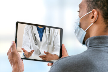 view of a afro-american young man with aCovid 19 proctection mask having an online consultation with his doctor