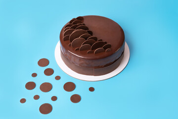Dark chocolate mousse cake covered with mirror glaze and decorated with round chocolate chips on blue background. Modern stylish cake. Close-up. Copy space.