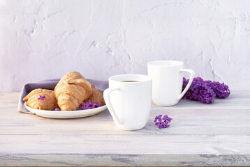 Fototapeta na wymiar Two beautiful porcelain cups of coffee with milk with croissants decorated with lilac flowers on white wooden table. Perfect breakfast concept. Copy space.