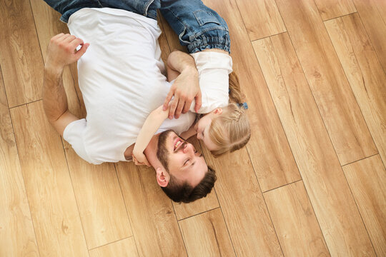 Top view of handsome young father and his cute little daughter looking at camera and smiling, lying on wooden floor. Hug and play. Father's day.