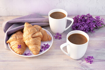 Two beautiful porcelain cups of coffee with milk with croissants decorated with lilac flowers on white wooden table. Perfect breakfast concept. Copy space.