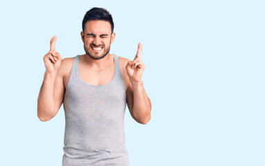 Young handsome man wearing swimwear and sleeveless t-shirt gesturing finger crossed smiling with...