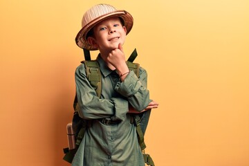 Cute blond kid wearing explorer hat and backpack smiling looking confident at the camera with crossed arms and hand on chin. thinking positive. - obrazy, fototapety, plakaty