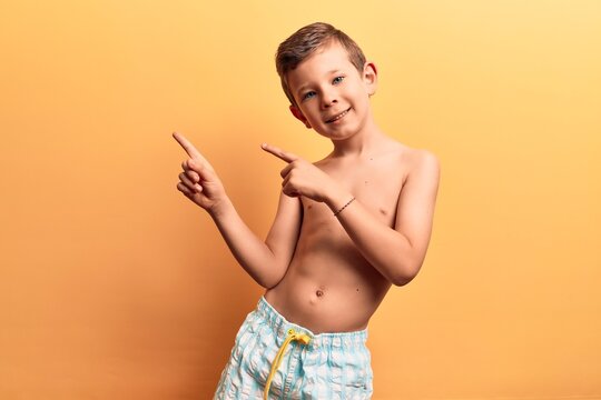 Cute blond kid wearing swimwear smiling and looking at the camera pointing with two hands and fingers to the side.