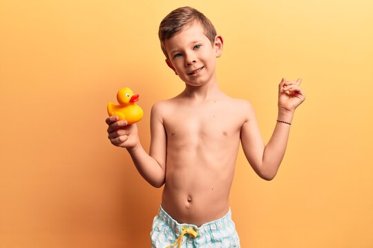 Cute blond kid wearing swimwear holding duck toy smiling happy pointing with hand and finger to the side