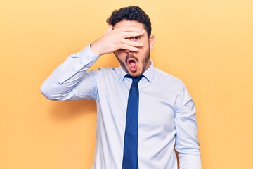 Young hispanic man wearing business clothes peeking in shock covering face and eyes with hand, looking through fingers afraid