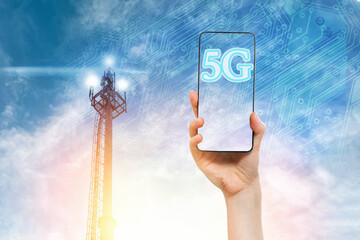 A woman's hand holds a smartphone against a blue sky with a 5G telecommunications tower. Concept of...