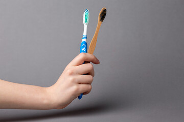 Oral hygiene. A female hand holds bamboo and plastic toothbrushes in her hand. Gray background. The concept of choosing eco-products. Copy space