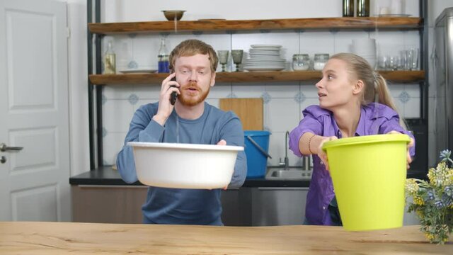 Young couple with pots collecting water falling from ceiling and calling plumber