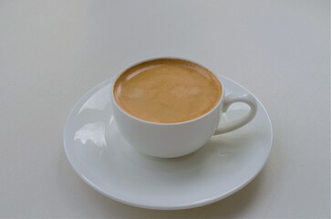 A small white cup with aromatic light brown coffee on a saucer, on a light background