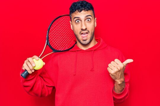 Young hispanic man playing tennis holding racket and ball pointing thumb up to the side smiling happy with open mouth