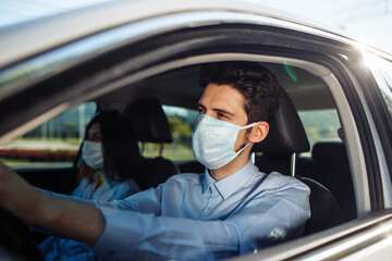 Young man taxi driver wears sterile medical mask in the car. A boy behind the car steering wheel safely driving for business matter keeping social distance. Coronavirus pandemic concept.