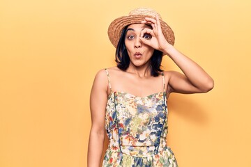 Young beautiful latin woman wearing summer hat doing ok gesture shocked with surprised face, eye looking through fingers. unbelieving expression.