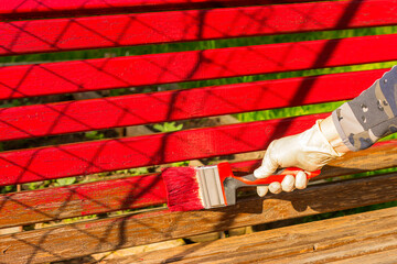 Painting with a brush a country bench on a garden site with bright red paint.