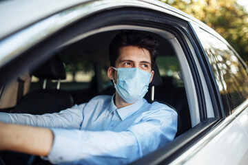 Fototapeta na wymiar Young man sits behind the steering wheel in the car wearing sterile medical mask. Boy taxi driver works hard during coronavirus outbreak. Social distance, virus spread prevention and treat concept.
