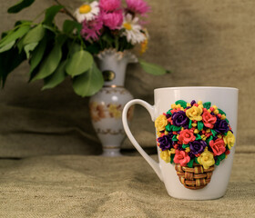 The original mug with decorative figurines made with their own hands. Figures on the cup are fashioned hand made from colored clay. Vase with fresh flowers on a background. High quality photo
