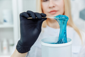 Soft blue wax is lifted with a wooden spatula from a white tube. The spatula is held by a hand in a...
