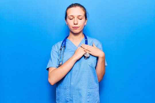 Young blonde woman wearing doctor uniform and stethoscope smiling with hands on chest with closed eyes and grateful gesture on face. health concept.