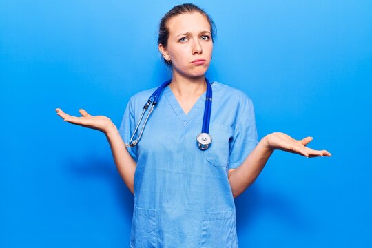 Young blonde woman wearing doctor uniform and stethoscope clueless and confused expression with arms and hands raised. doubt concept.