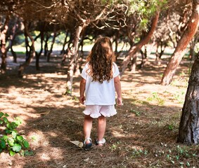 Adorable blonde child on back view standing around trees on the park