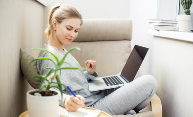 Woman in gray home suit with laptop in her hands sitting in chair on apartment