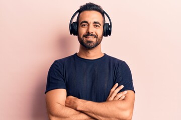Young hispanic man listening to music using headphones happy face smiling with crossed arms looking...