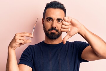 Young hispanic man holding syringe with angry face, negative sign showing dislike with thumbs down,...