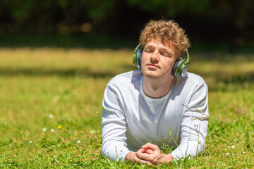 young red-haired man in headphones lies on the green grass enjoying music on a sunny day