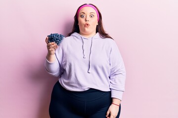 Young plus size woman holding blueberries scared and amazed with open mouth for surprise, disbelief...